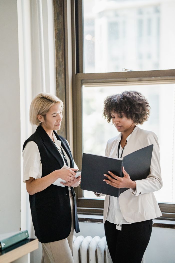 The Art of Drawing Readers In: Your attractive post title goes here Two women in business attire discussing documents by a window in the office.